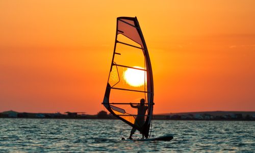 Seascape of still sea surface, man practicing wind surfing and golden sunset in sky on summer clear day. Still landscapes of travels and destination scenics